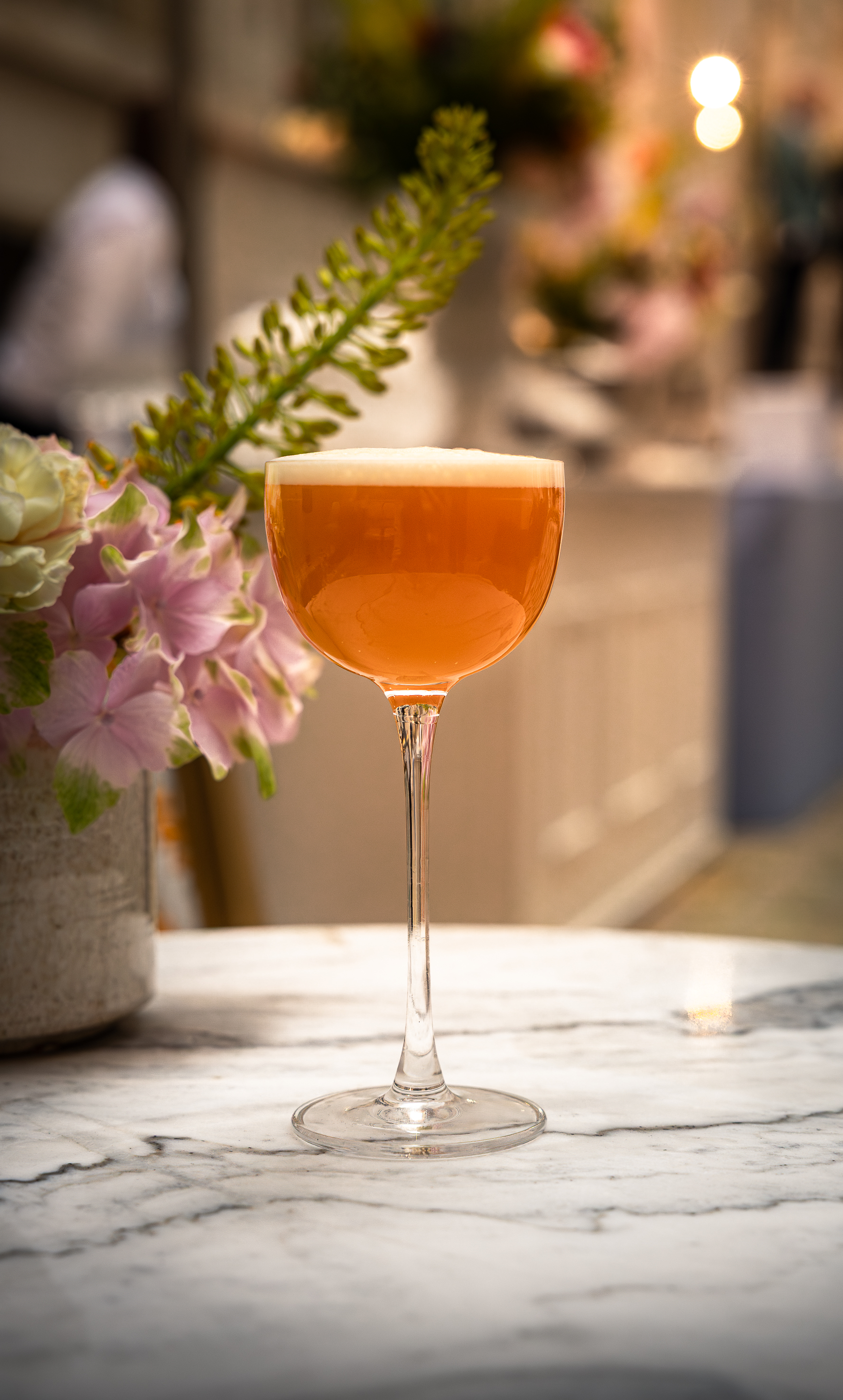 Cocktail ambré à la mousse fine dans un verre sur pied, sur une table en marbre près de fleurs