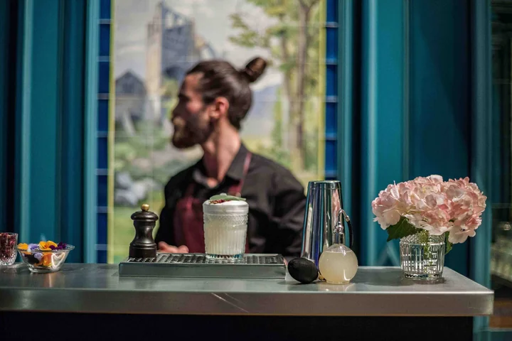 Barman au chignon derrière le bar, décor bleu canard et grande fenêtre, bouquet de pivoines roses sur le comptoir