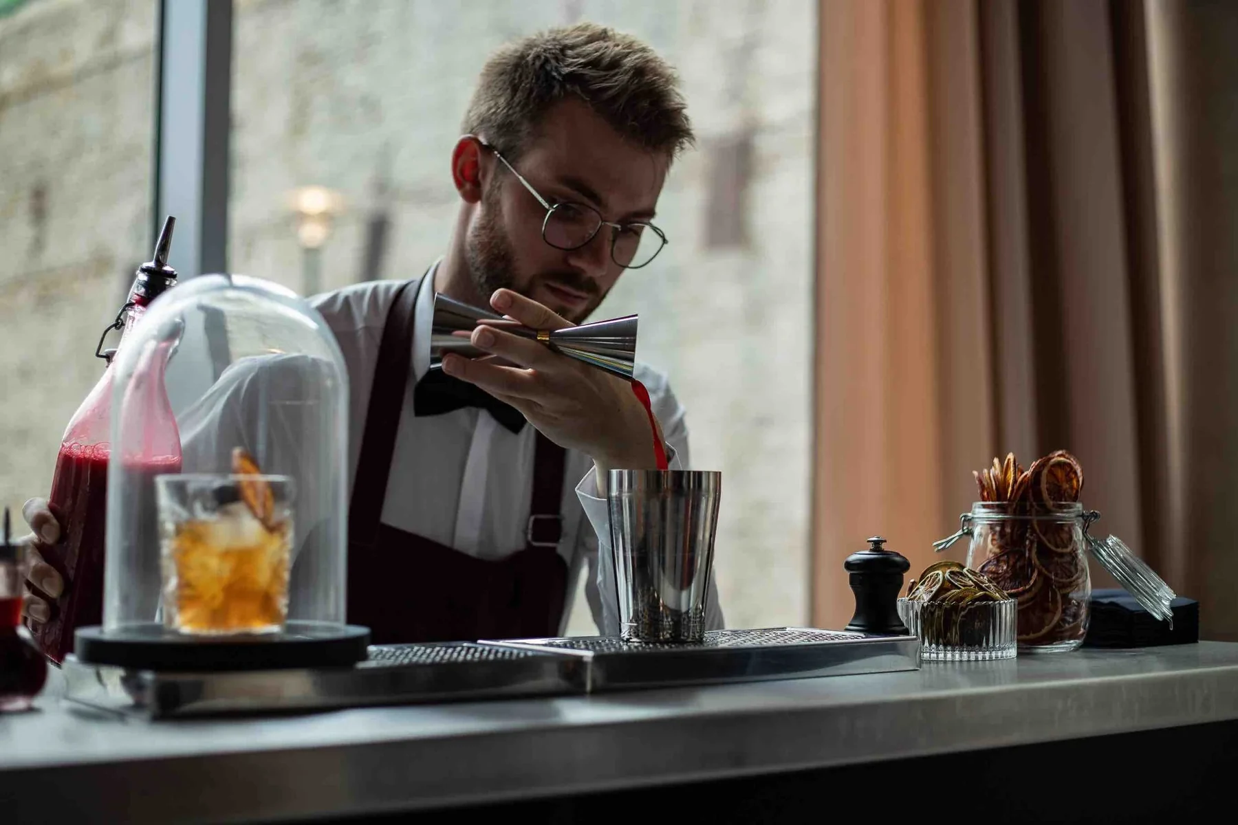 Barman à lunettes et nœud papillon devant une cloche de fumage, cocktail ambré sur le bar près d'une baie vitrée