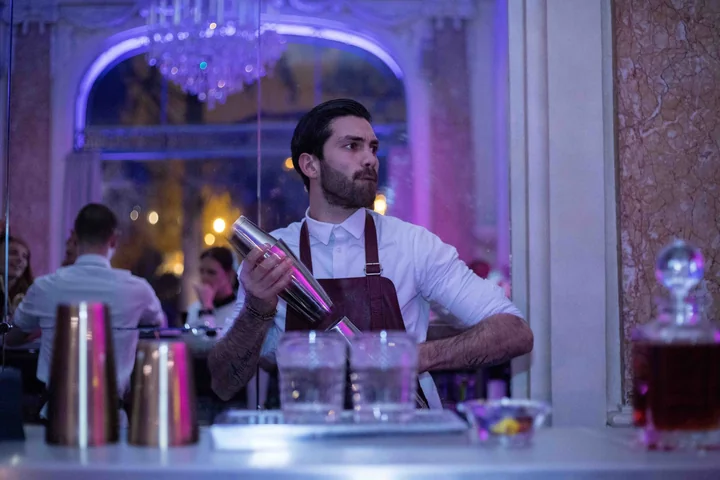Barman secouant un shaker dans une salle de bal éclairée de violet, lustres et invités attablés en arrière-plan