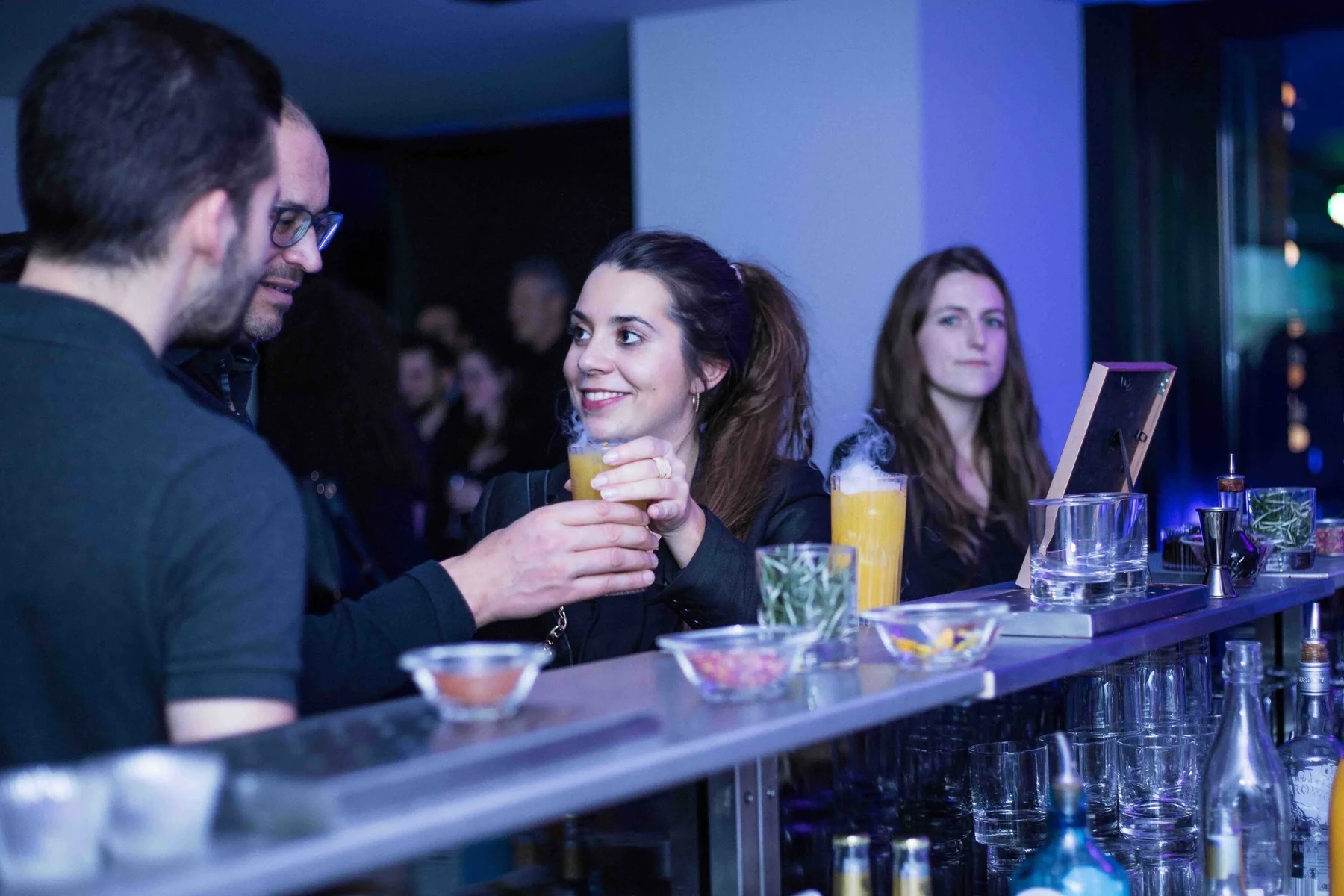 Invités accoudés au bar échangeant avec un barman lors d'une soirée, cocktails jaunes sur le comptoir