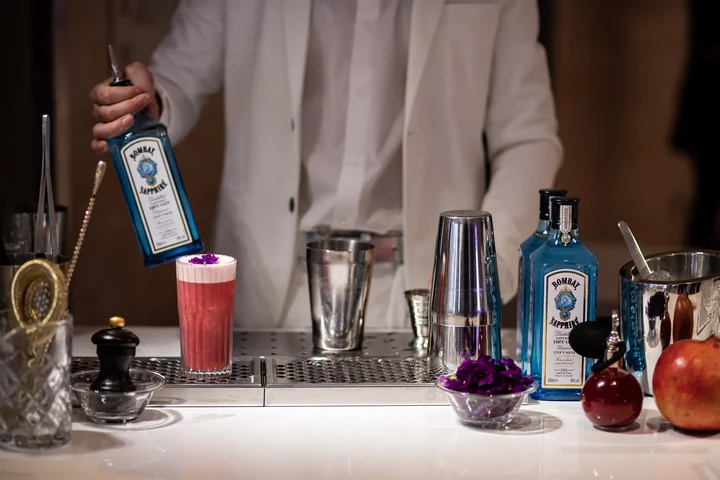Barman en veste blanche au bar à gin : bouteilles bleues, cocktail rose, fleurs violettes et grenades