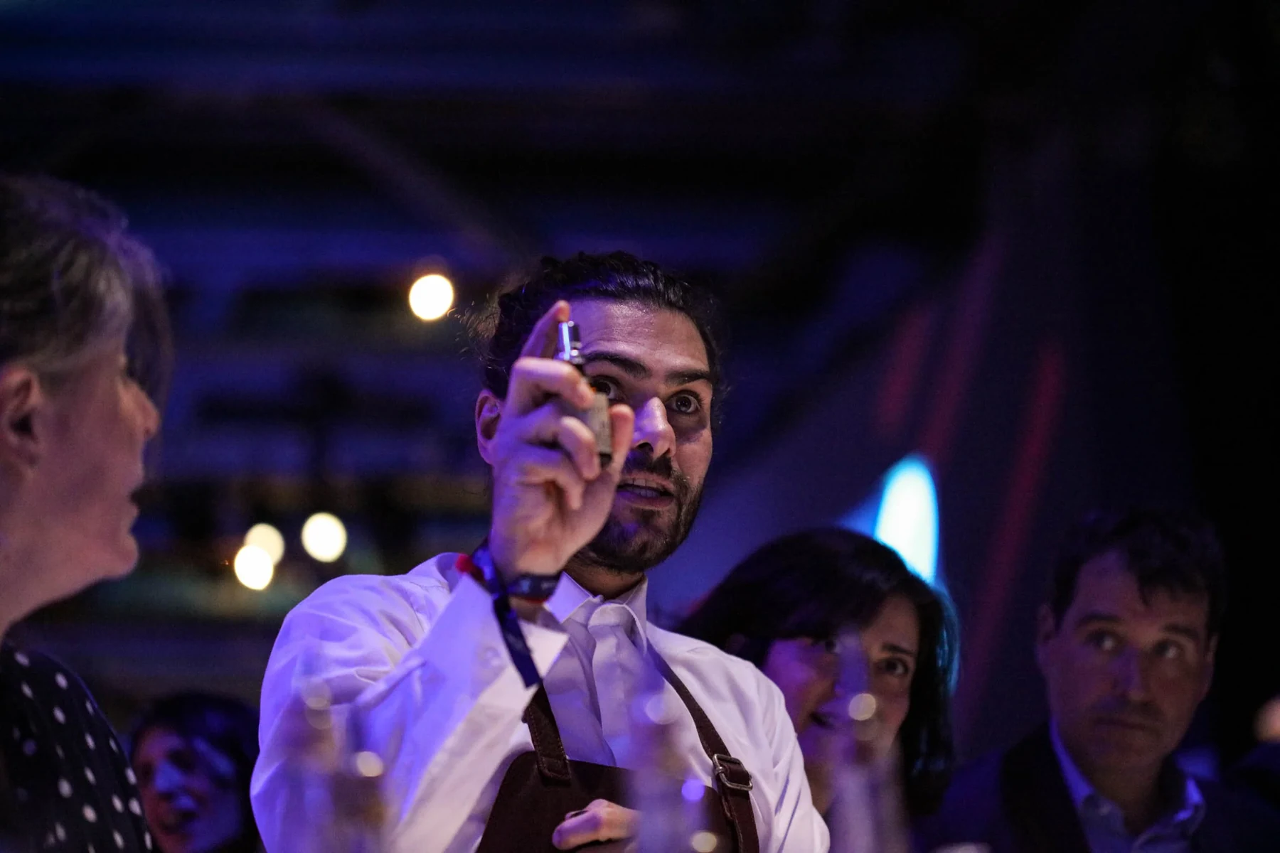 Barman en tablier levant un petit instrument de bar au milieu des invités, soirée aux lumières bleues