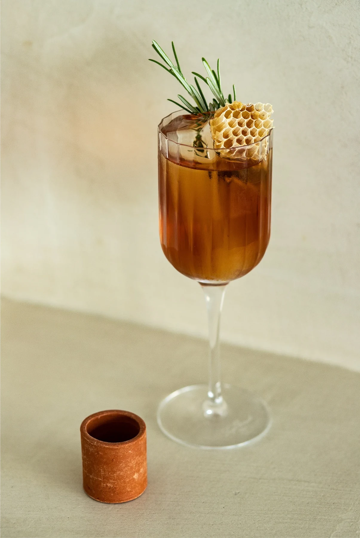 Cocktail ambré en verre à pied garni de romarin et d'une tranche d'agrume déshydratée, sur fond crème