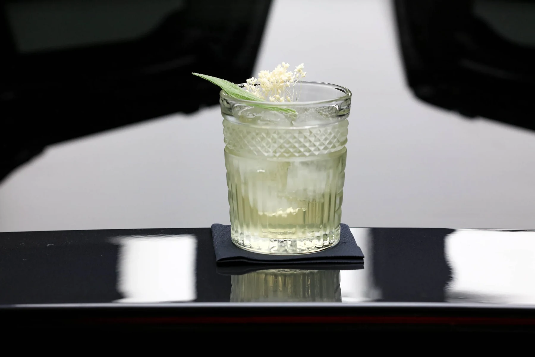 Cocktail clair en verre ciselé garni de fleurs blanches, posé sur un sous-verre noir