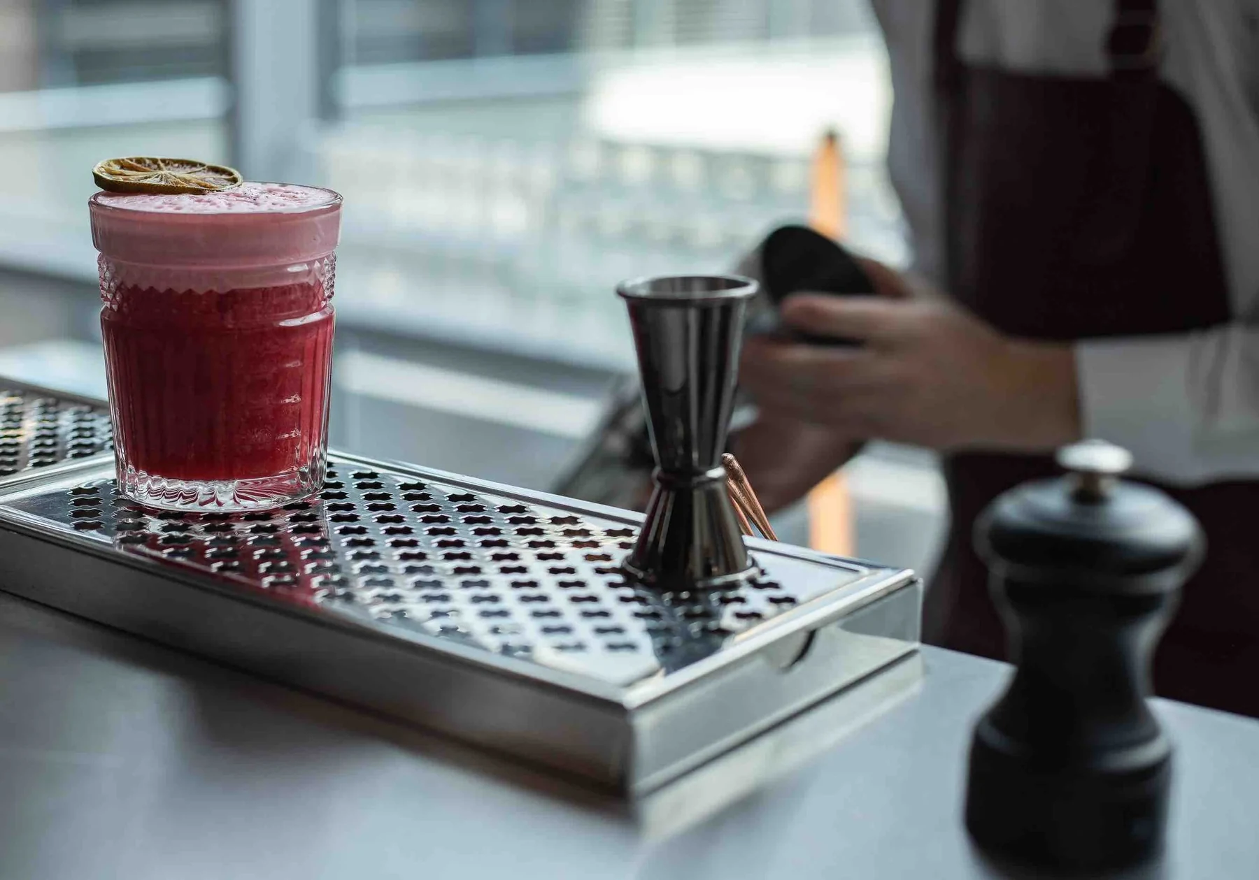 Cocktail rouge mousseux sur égouttoir inox, à côté d'un jigger et d'un moulin noir