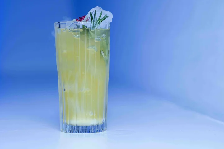 Grand verre de cocktail jaune garni de fleurs et d'herbes, sur fond bleu dégradé