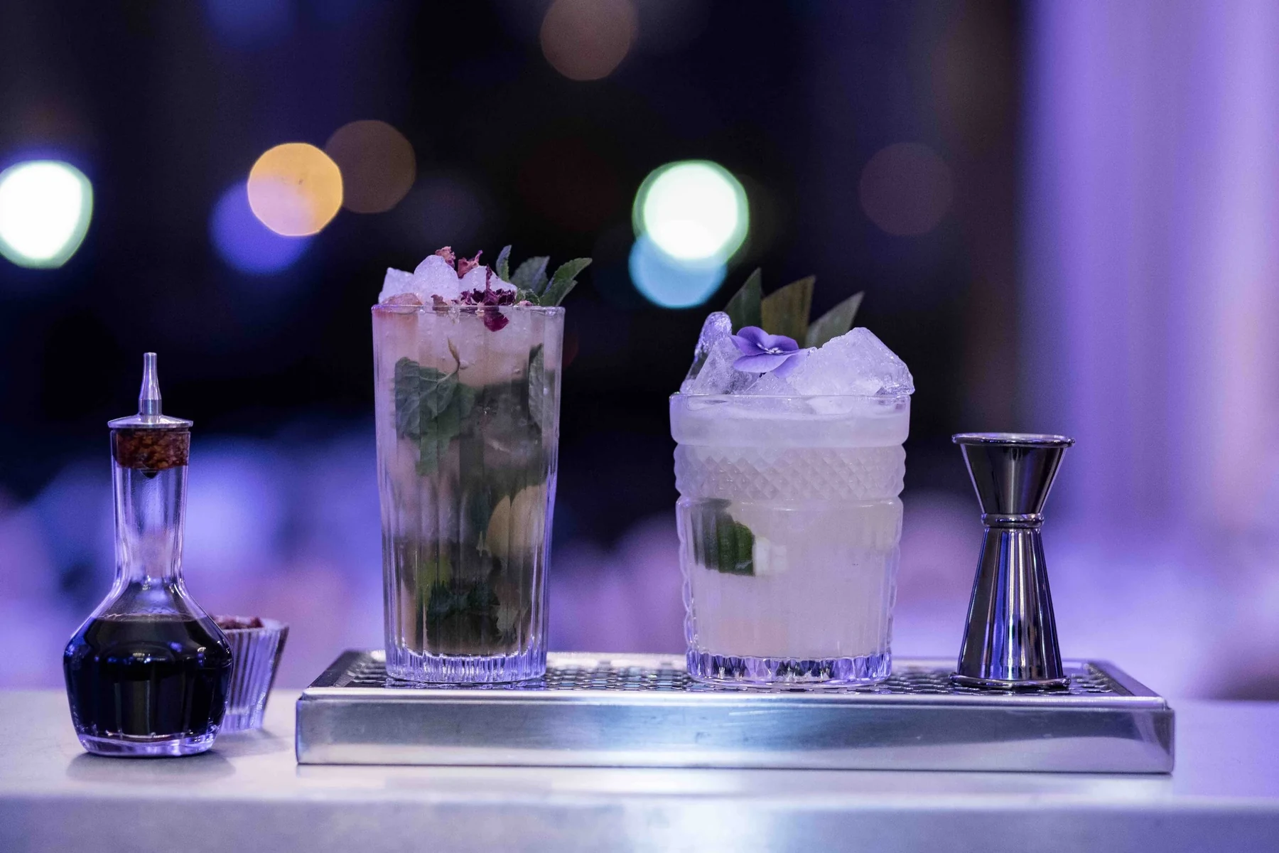 Deux cocktails glacés garnis de fleurs, jigger et bouteille sur plateau inox, bokeh violet