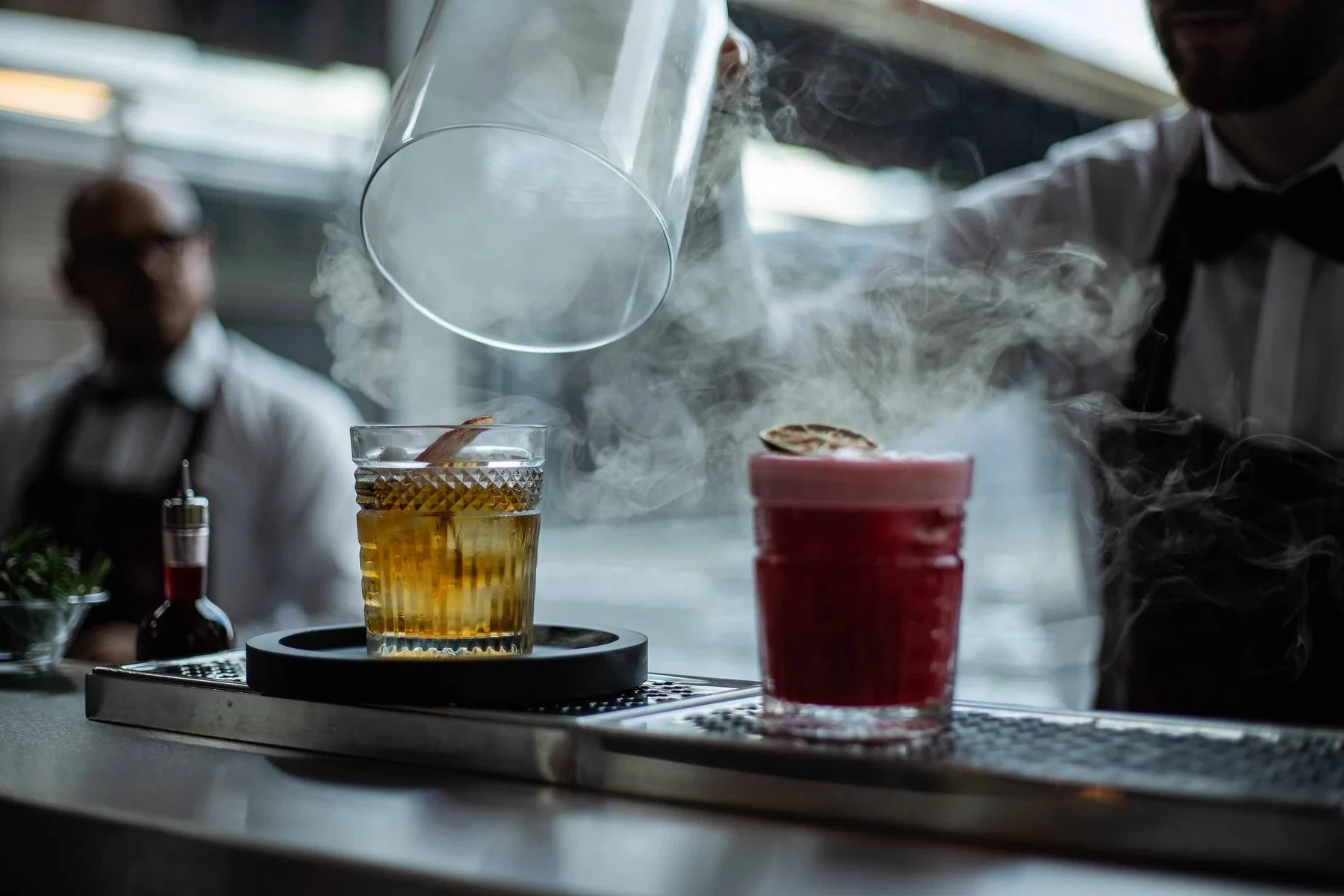 Cloche de fumage soulevée au-dessus d'un old fashioned ambré, cocktail rouge à côté