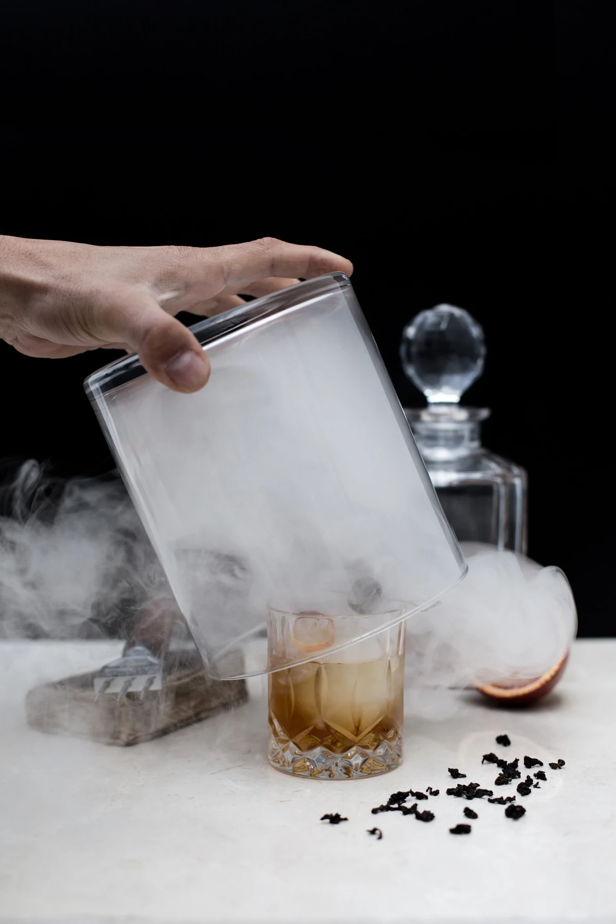Main soulevant une cloche de verre libérant la fumée d'un cocktail sur fond sombre
