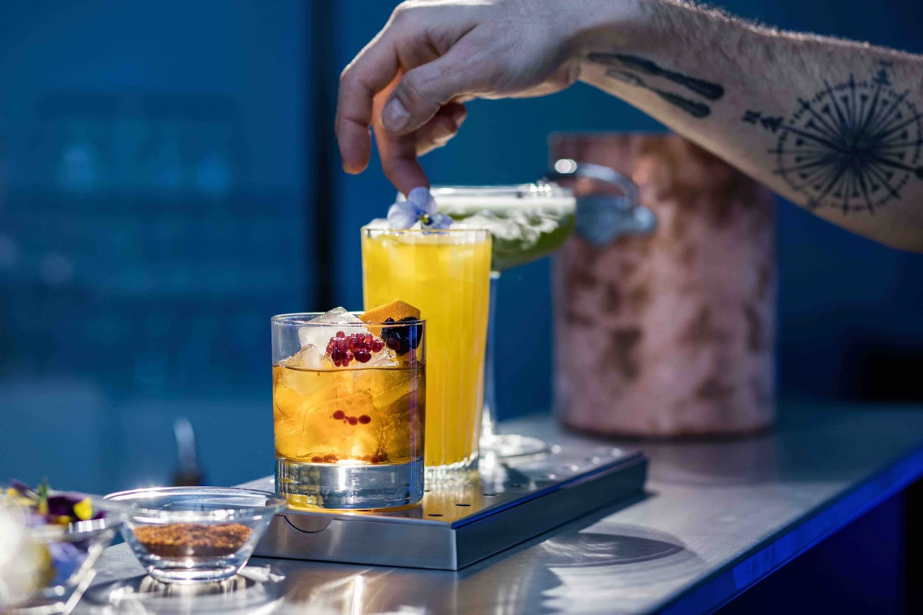 Main tatouée déposant une garniture sur un cocktail jaune en verre haut, second cocktail garni de fruits rouges, lumière bleue
