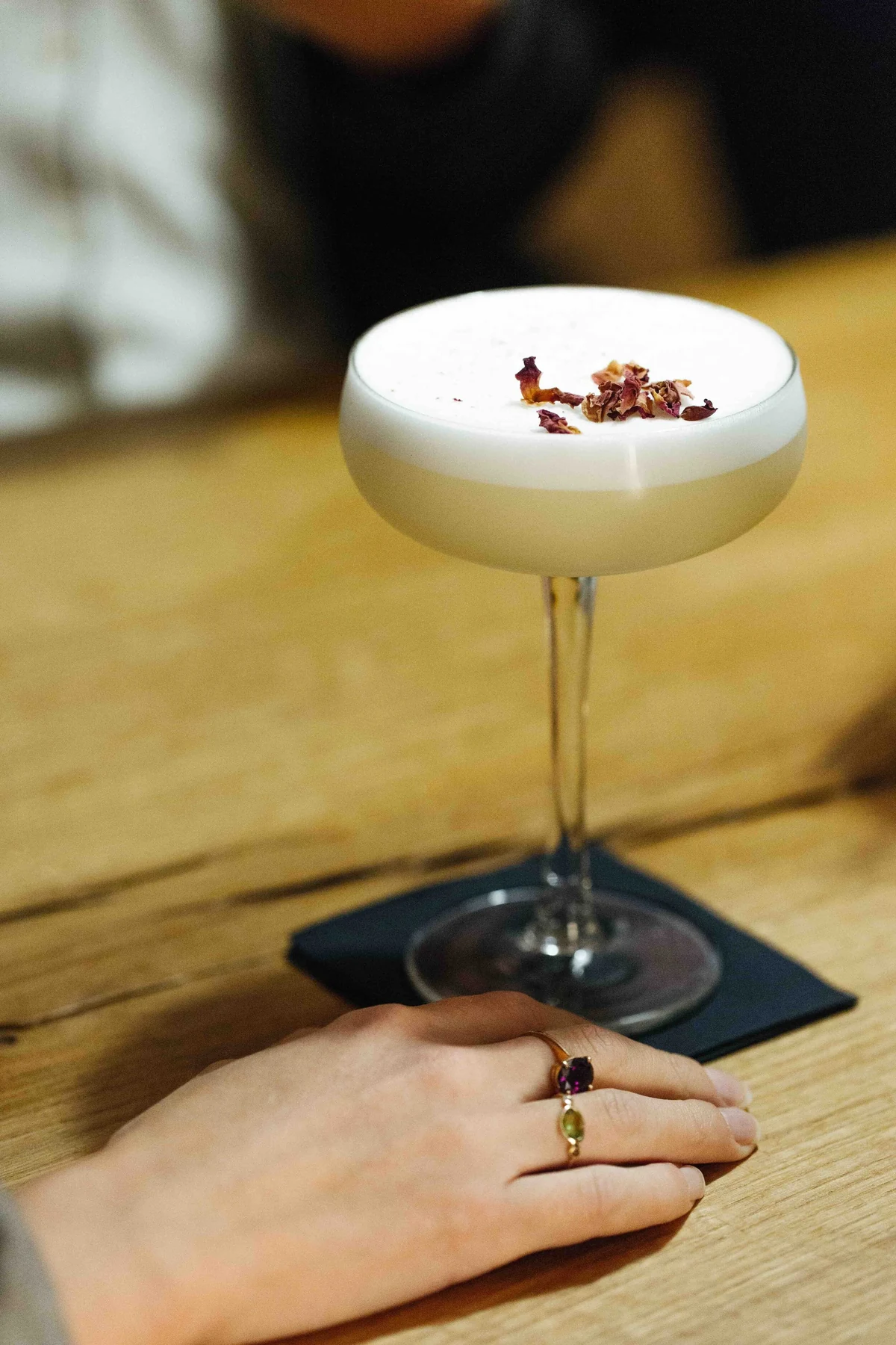 Main tenant une coupe de cocktail à mousse blanche garnie de pétales, sur une table en bois