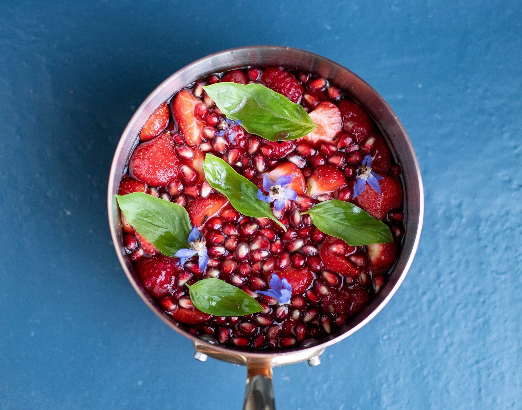 Vue plongeante d'une casserole de fruits rouges, grenade, feuilles de basilic et fleurs violettes sur fond bleu